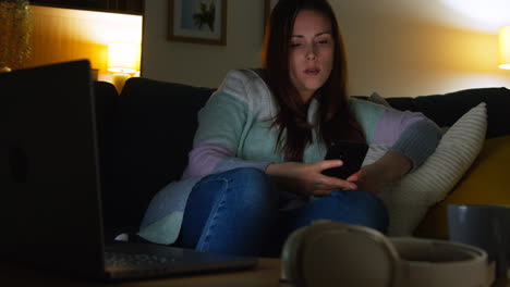 Mujer-Sentada-En-Un-Sofá-En-Casa-Por-La-Noche-Transmitiendo-O-Viendo-Películas-O-Programas-En-Una-Computadora-Portátil-Y-Navegando-Por-Internet-En-El-Teléfono-8